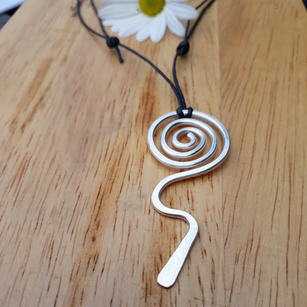 Silver spiral pendant on a black cord with a white flower in the background on a wooden surface