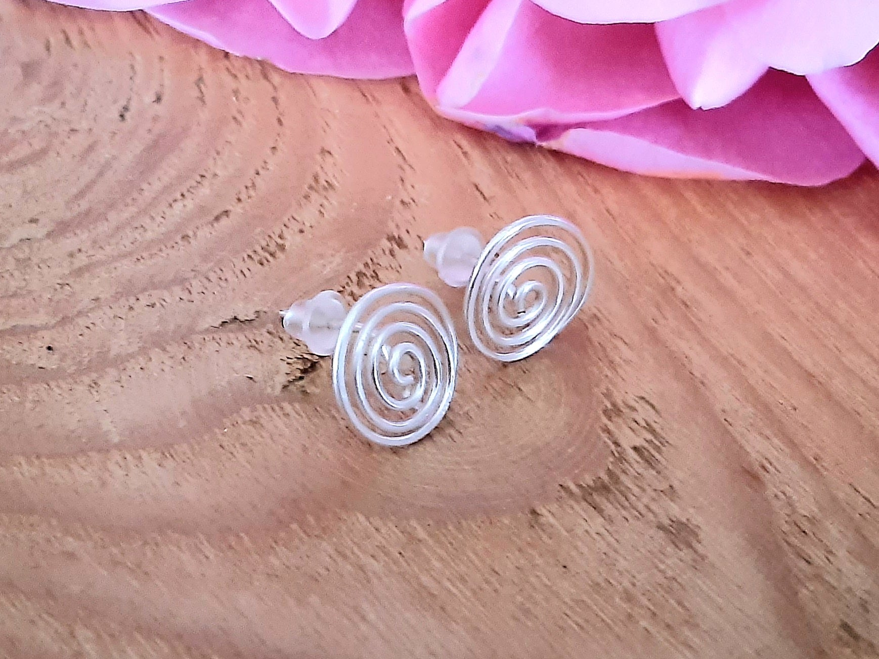 Silver spiral earrings on a wooden surface with pink flowers in the background