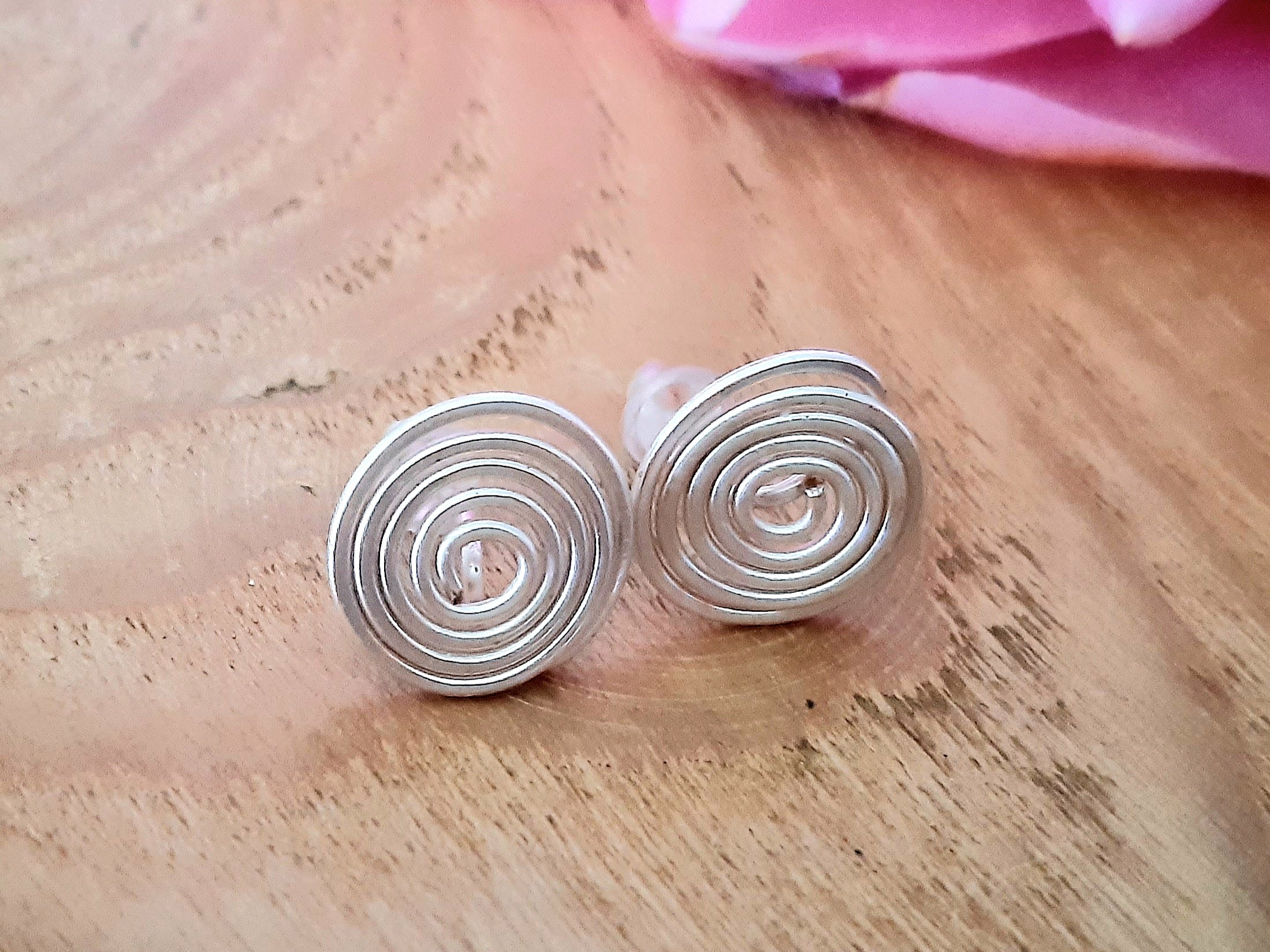 Silver spiral earrings on a wooden surface with a pink flower in the background