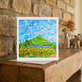 Colourful abstract artwork of Glastonbury Tor greeting card on a wooden surface with a stone wall background