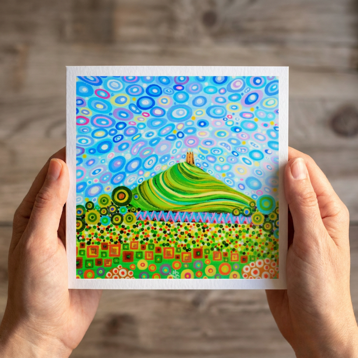 Colourful abstract artwork of Glastonbury Tor on a greeting card held by hands on a wooden surface