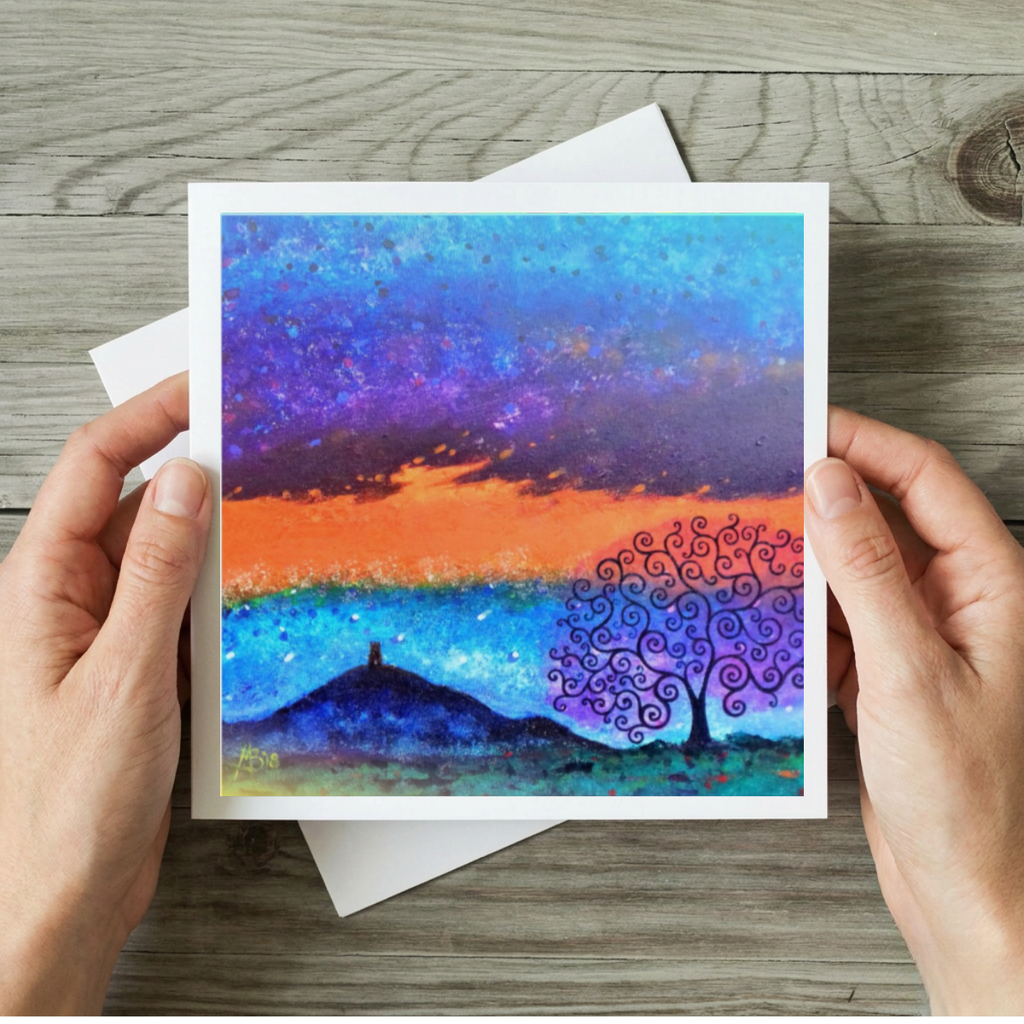 Artistic greeting card with a colourful tree and Glastonbury Tor design held by hands on a wooden surface