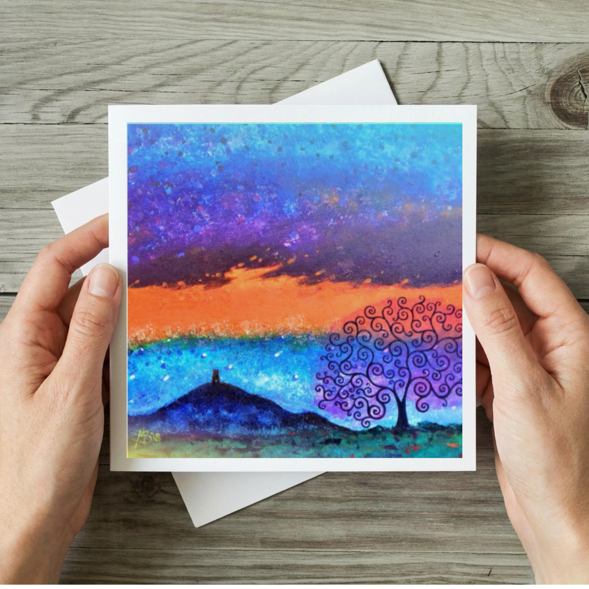 Artistic greeting card with a colourful tree and Glastonbury Tor design held by hands on a wooden surface