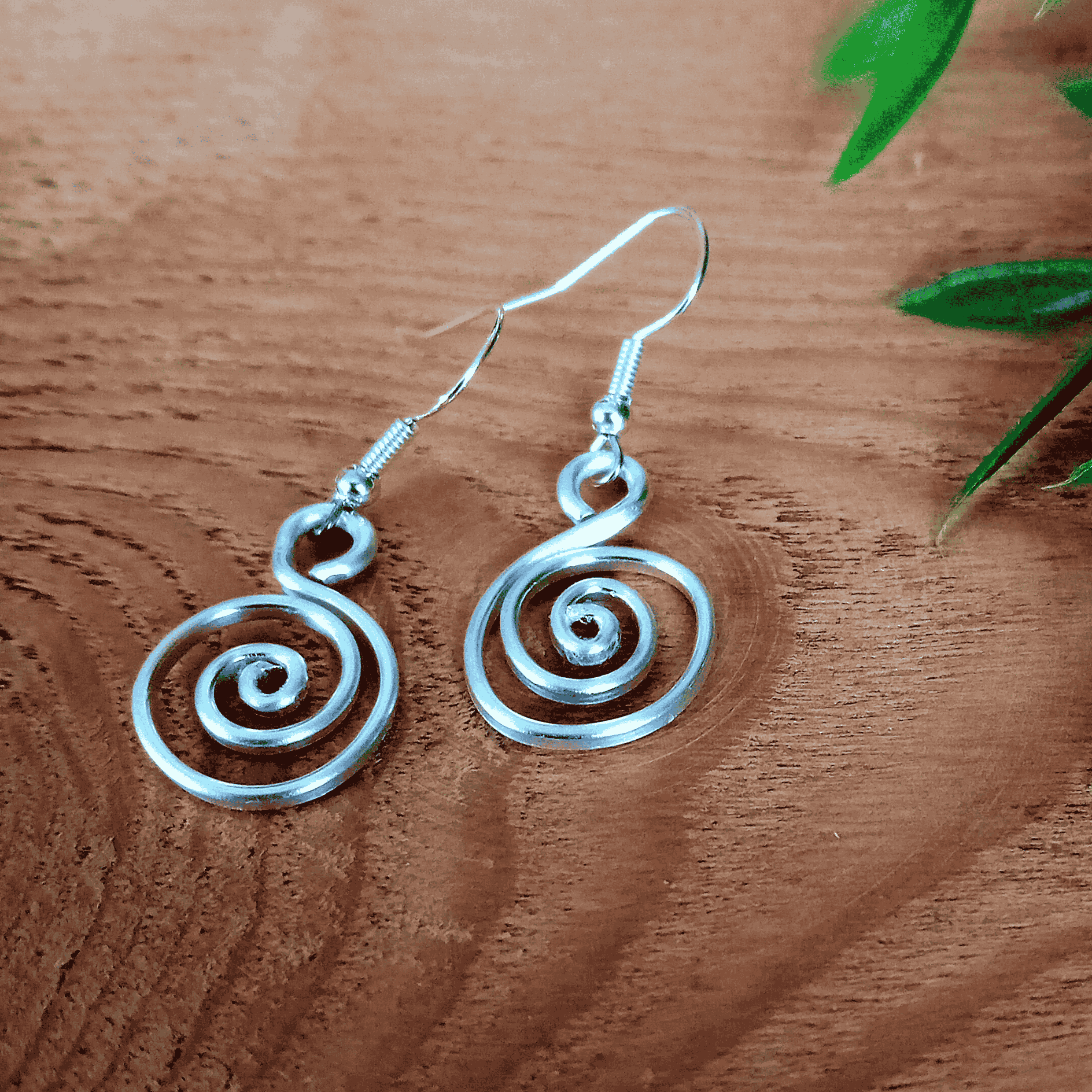 Silver spiral earrings on a wooden surface with green leaves.