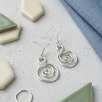 Silver spiral earrings on a textured surface with ceramic tiles and fabric.