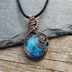 Blue Apetite stone pendant with copper wire wrapping on a black cord, placed on a wooden surface.
