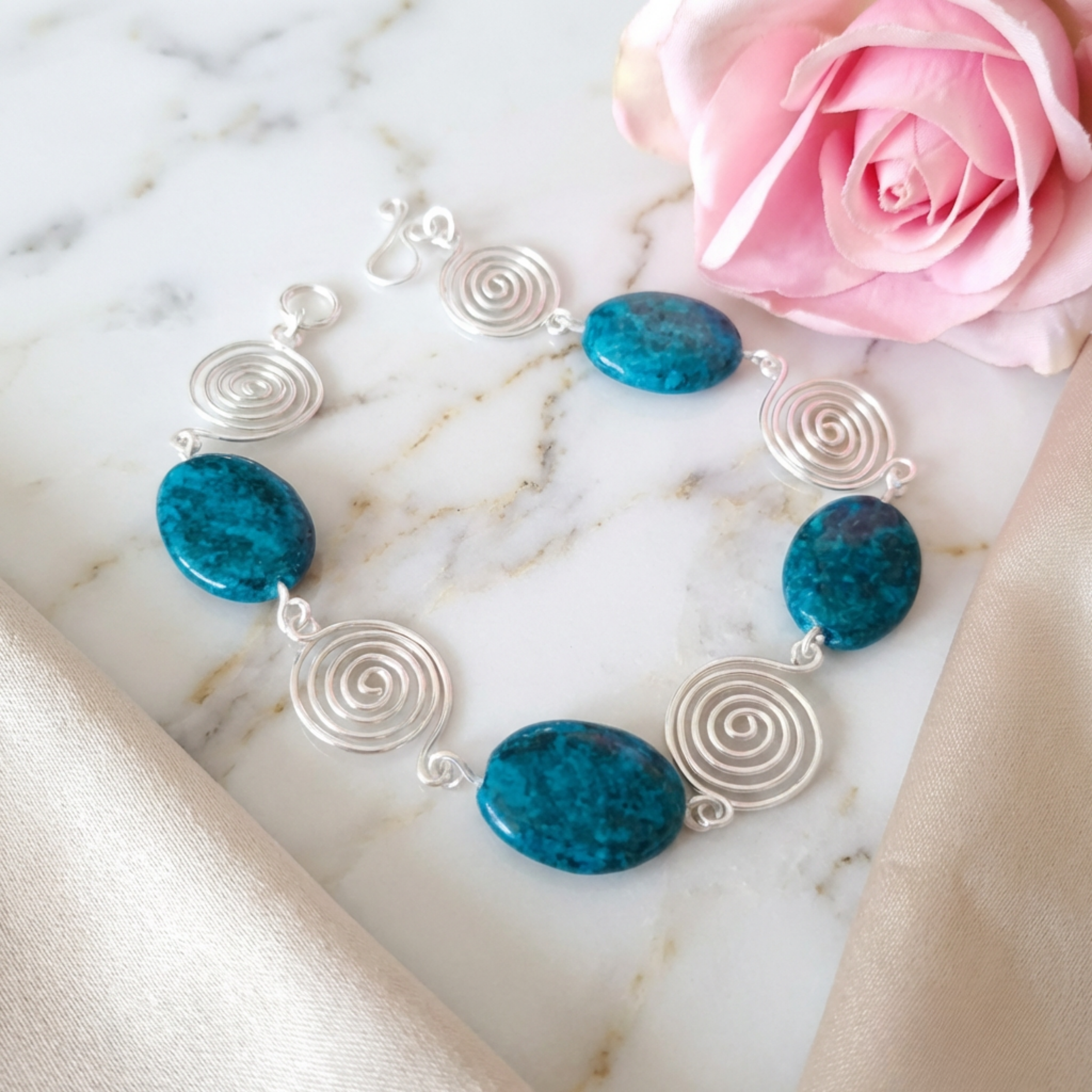 Bracelet with blue stones and silver spiral designs on a marble surface with a pink rose.