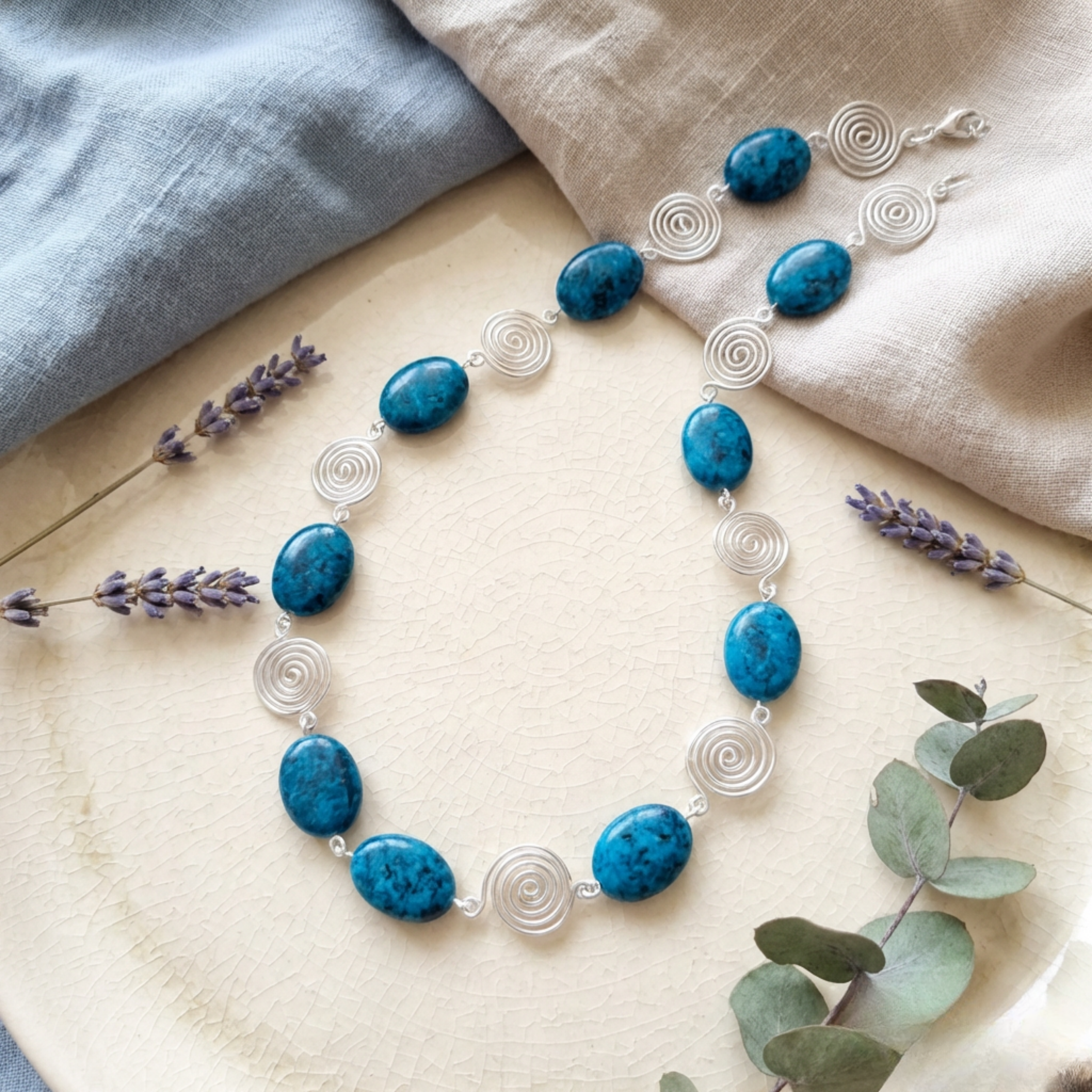 Necklace with blue stones and silver loops on a wooden surface with lavender and eucalyptus leaves.