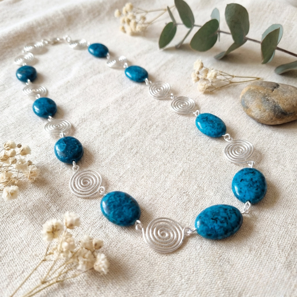 Necklace with blue beads and silver spiral accents on a textured surface with flowers and a stone.