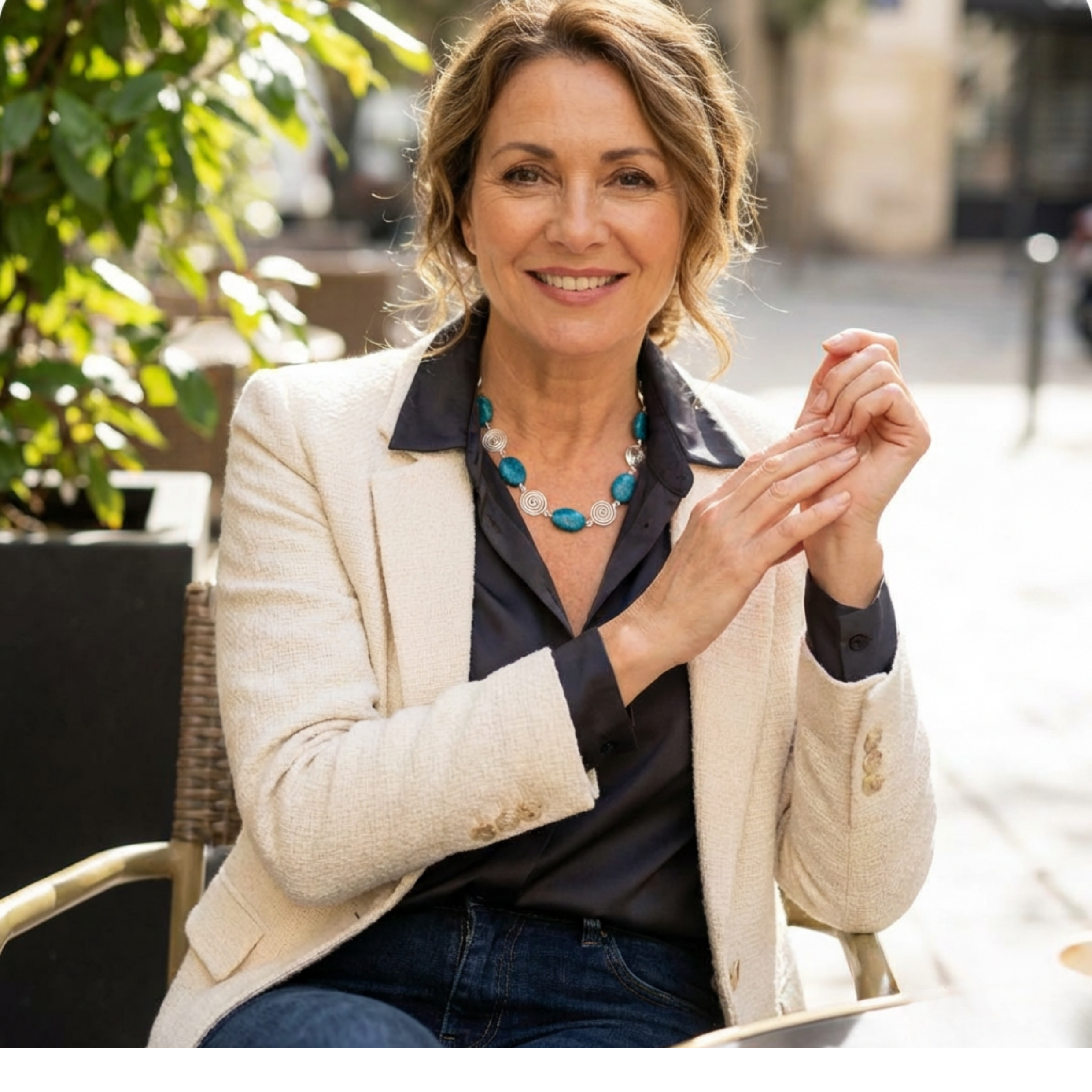 Woman sitting outdoors, smiling, wearing a beige blazer and blue jeans. Wearing a silver spiral and Azurite necklace