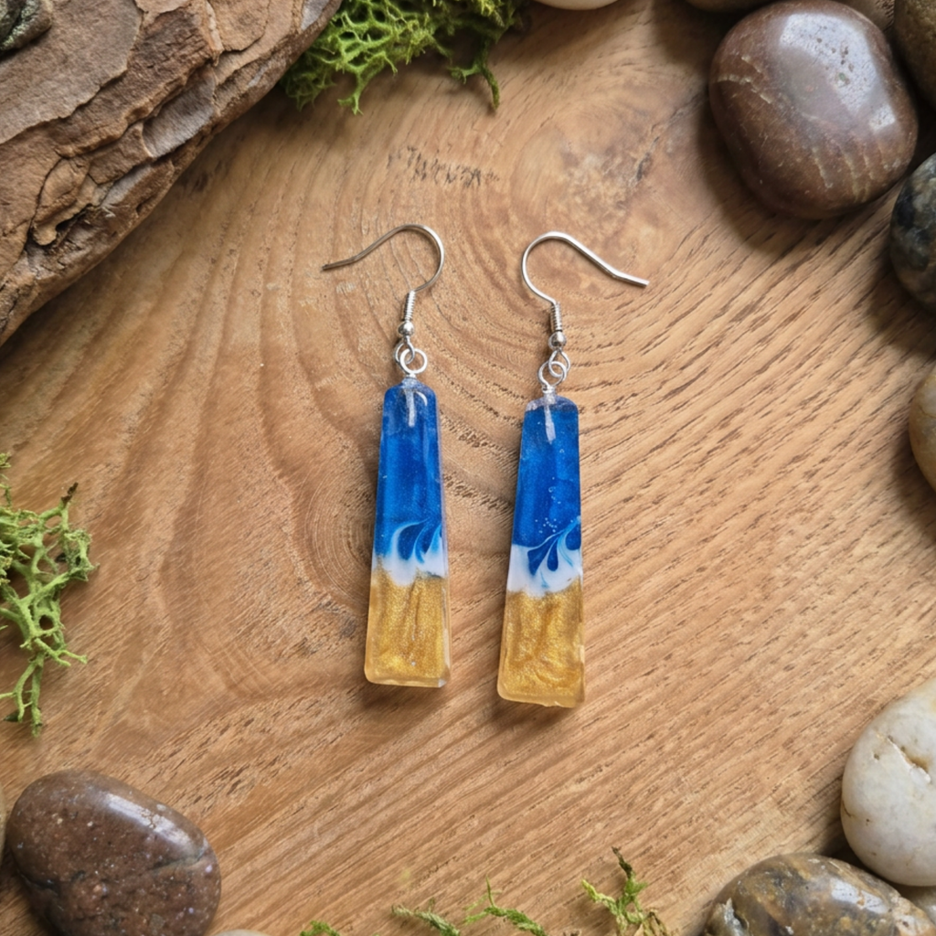 Pair of blue and gold earrings on a wooden surface with rocks and moss.