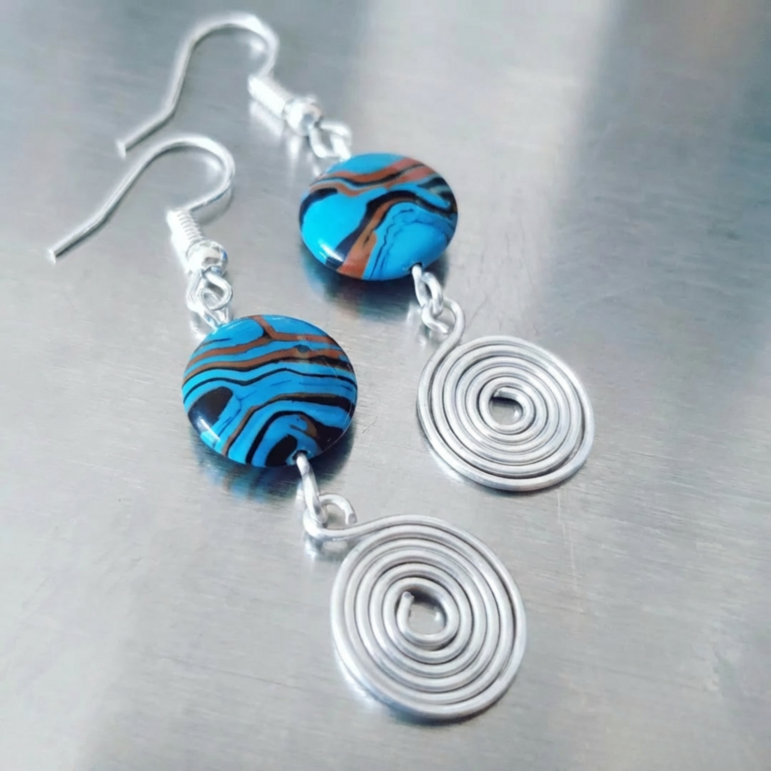 Earrings with blue and brown striped beads and silver spiral pendants on a metallic surface