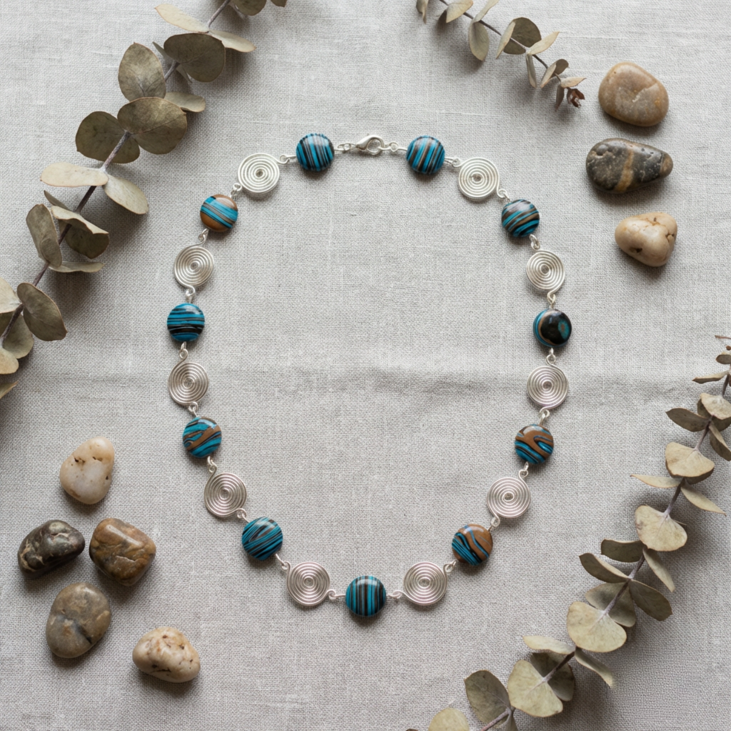 Necklace with blue and silver beads on a light gray surface with eucalyptus leaves and stones.
