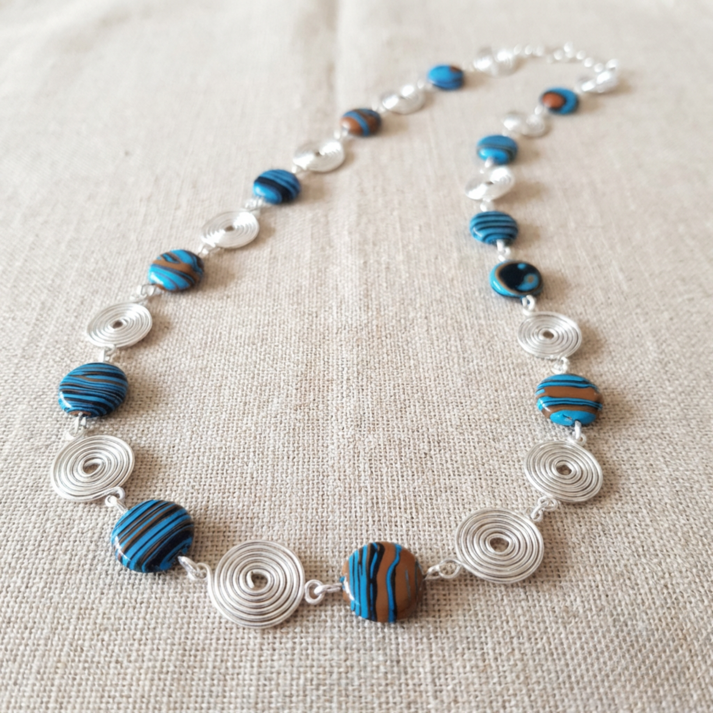 Necklace with blue and brown beads and silver spirals on a beige fabric background