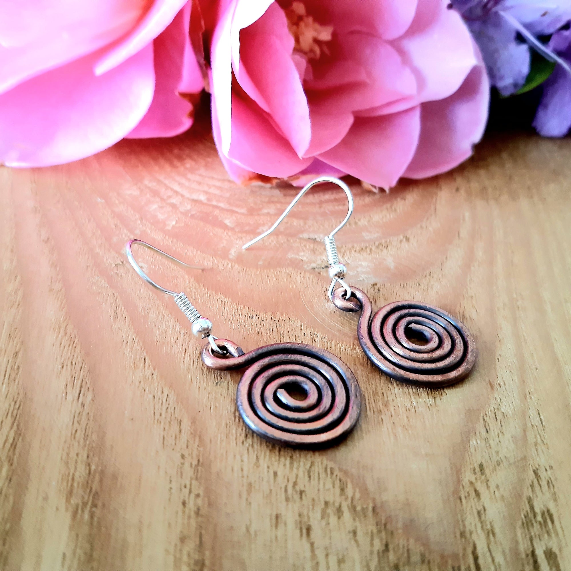 Copper spiral earrings on a wooden surface with pink flowers in the background