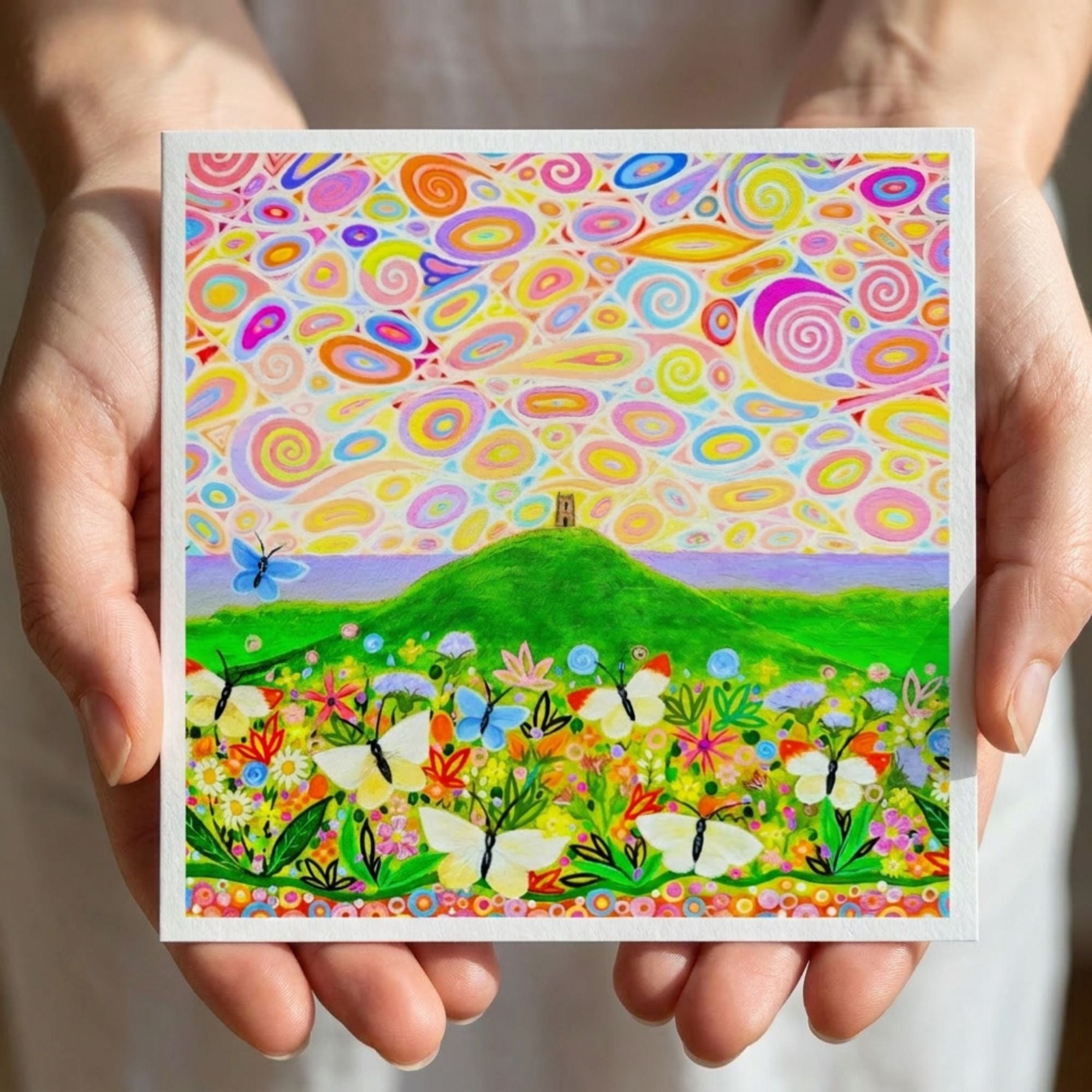 Colourful artwork of a landscape with flowers, butterflies, and Glastonbury Tor held by hands.