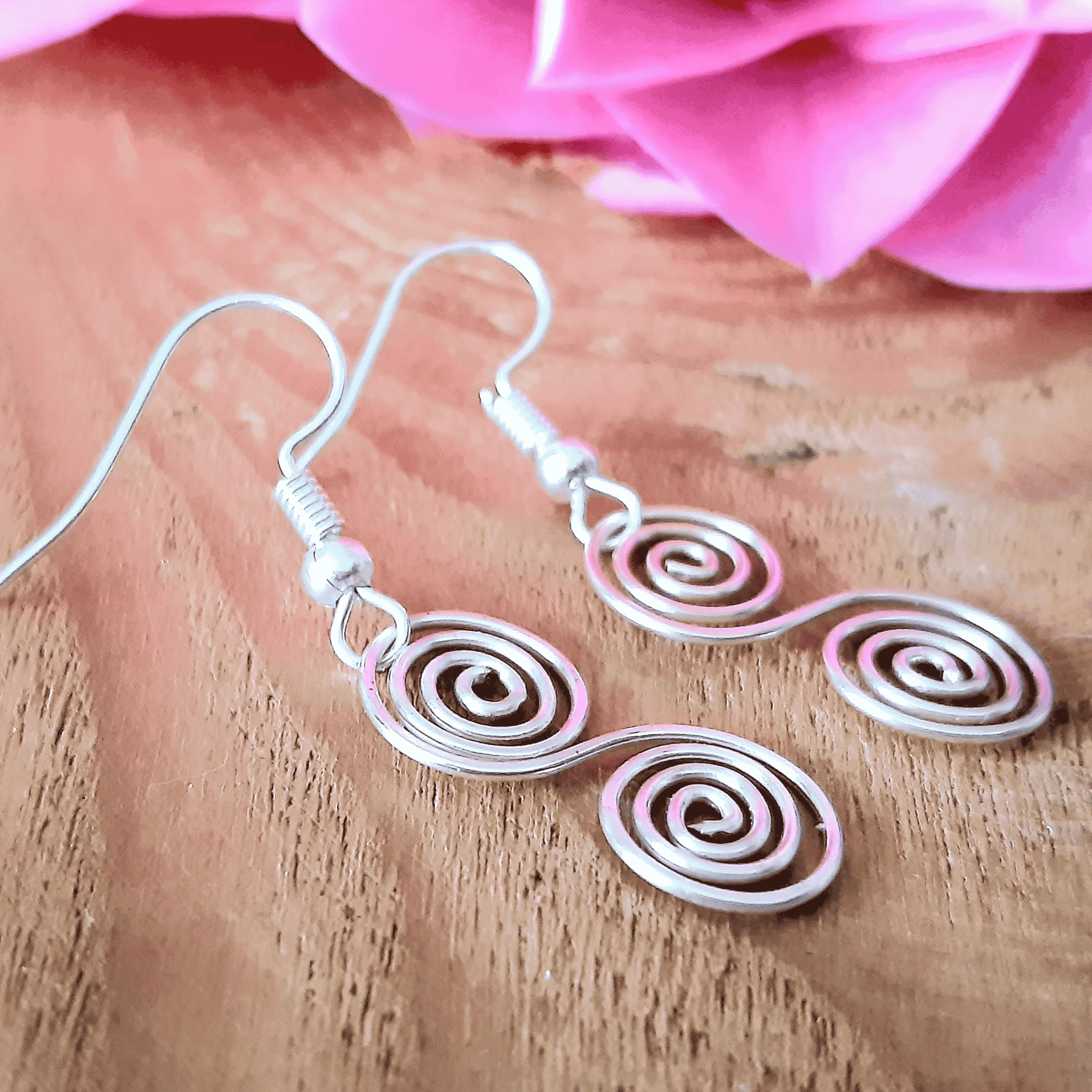 Silver spiral earrings on a wooden surface with pink flowers in the background
