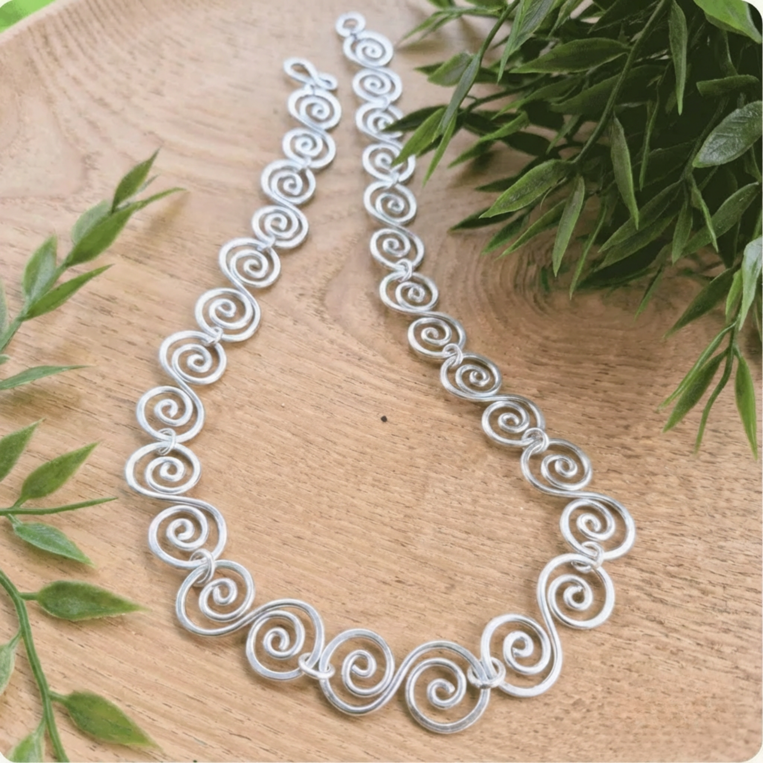 Silver necklace with intricate designs on a wooden surface with green leaves.