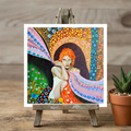Colourful artwork of a woman with red hair on a wooden easel with a potted plant in the background.