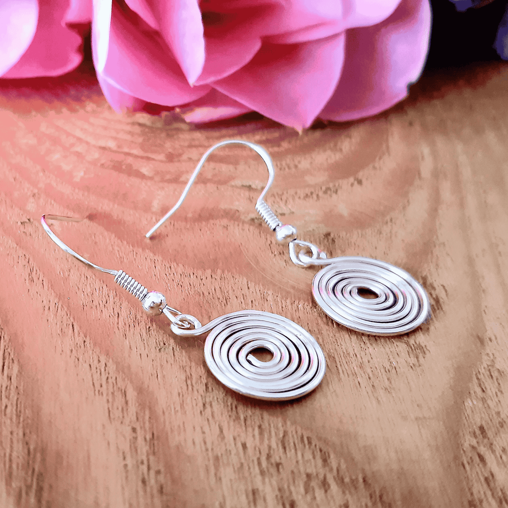 Silver spiral earrings on a wooden surface with pink flowers in the background