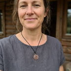 Woman wearing a gray shirt with a necklace featuring a spiral pendant, standing in front of a wooden cabin.