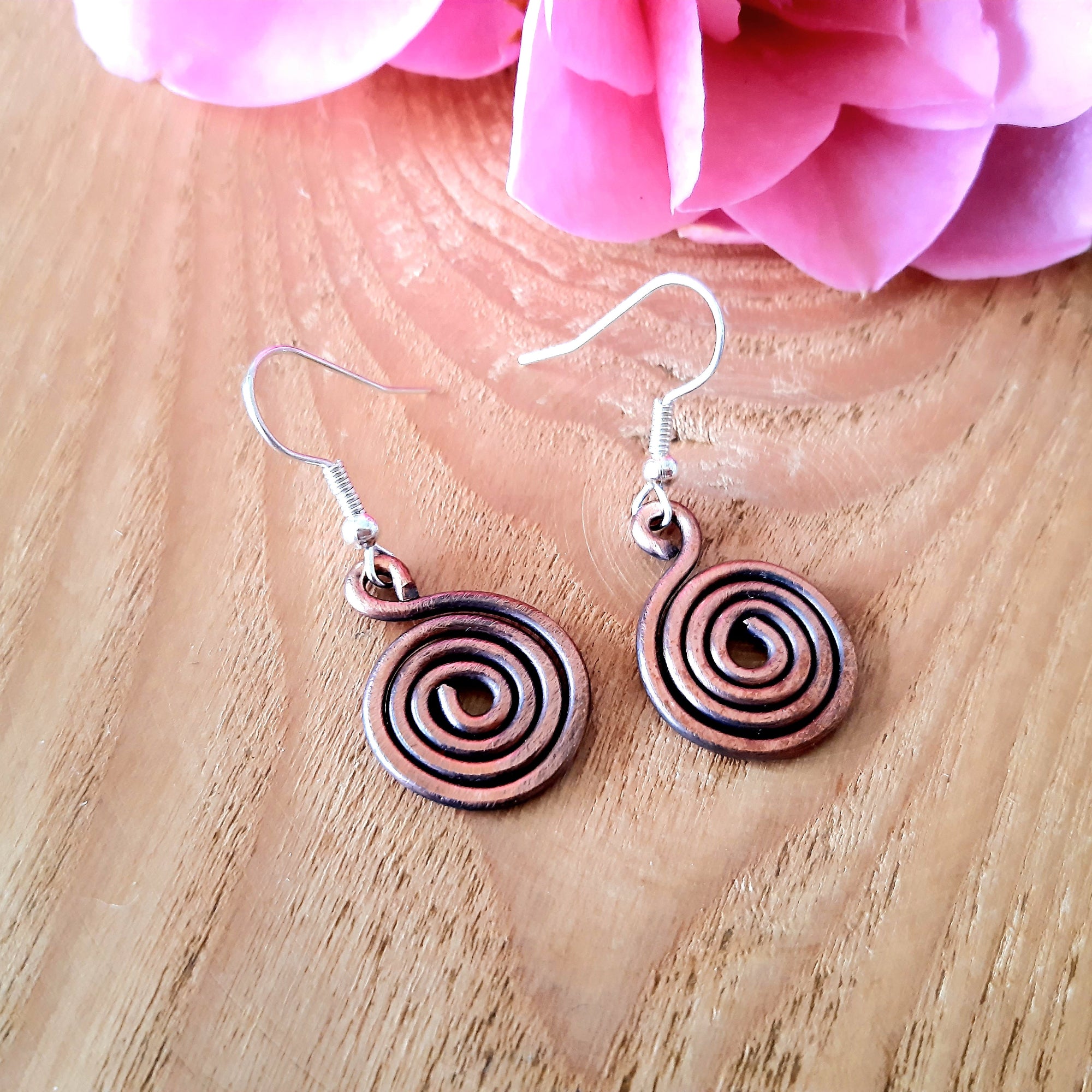 Copper spiral earrings on a wooden surface with pink flowers in the background