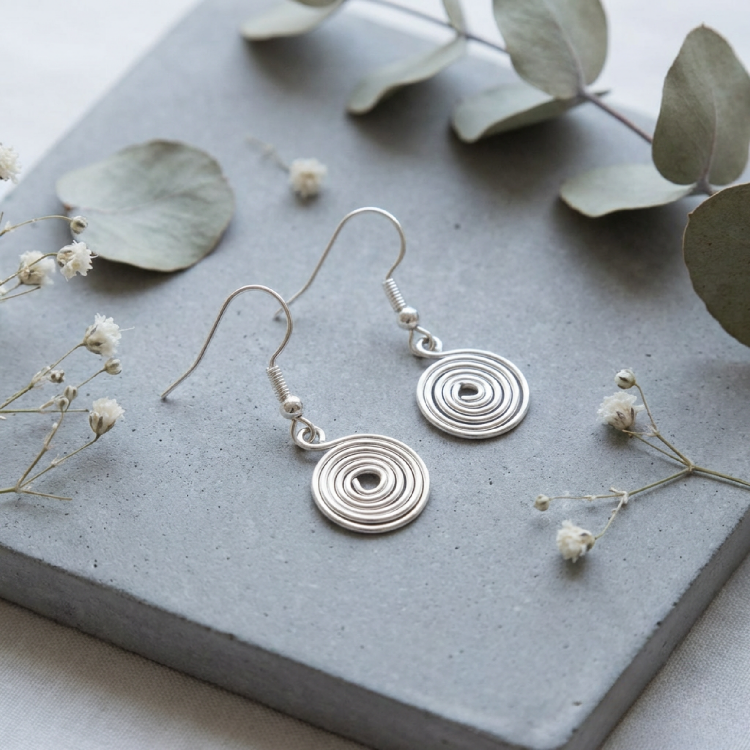 Silver circular spiral earrings on a grey surface with green leaves and small white flowers.