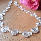 Silver necklace with circular links on a wooden surface with a pink flower in the background