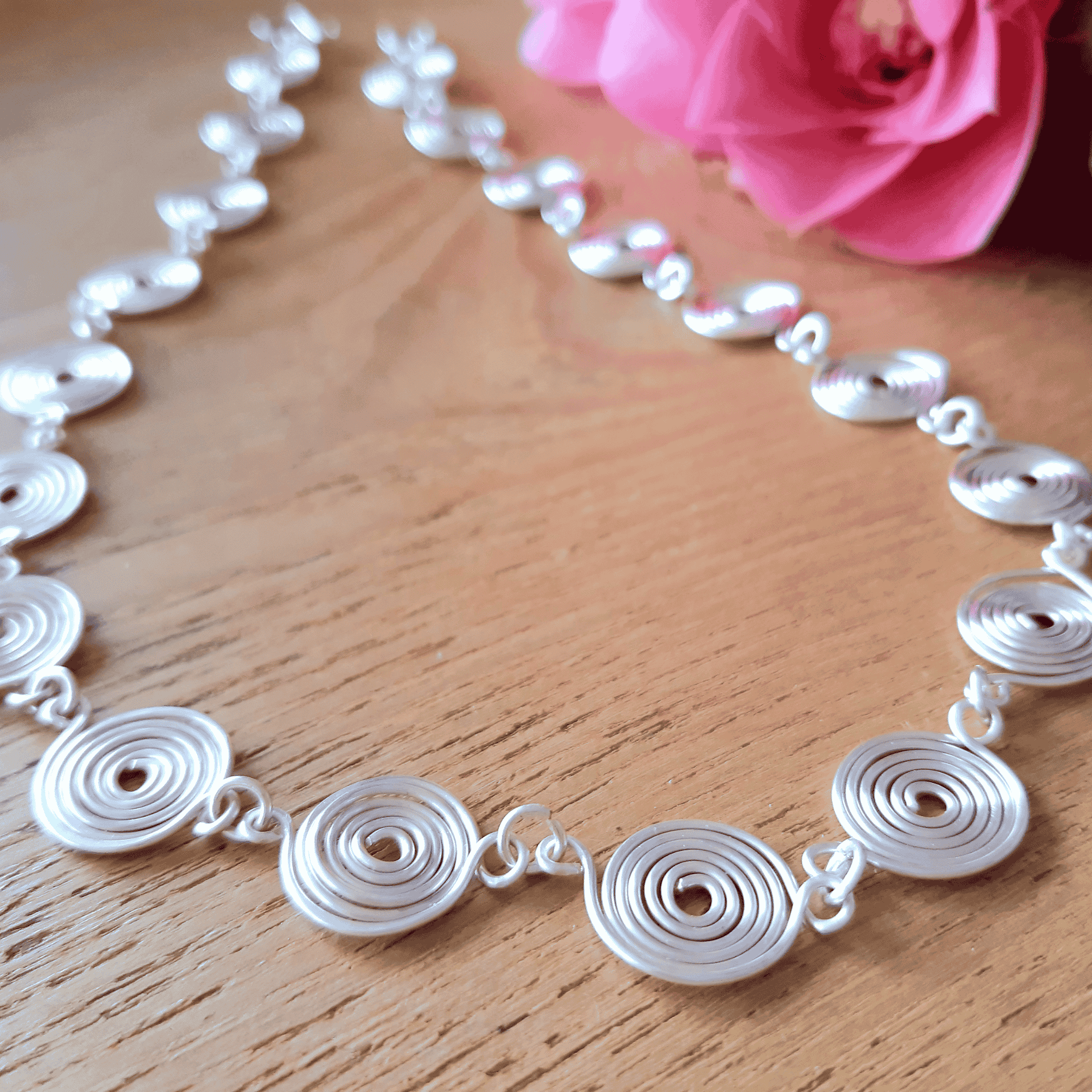 Silver necklace with circular links on a wooden surface with a pink flower in the background
