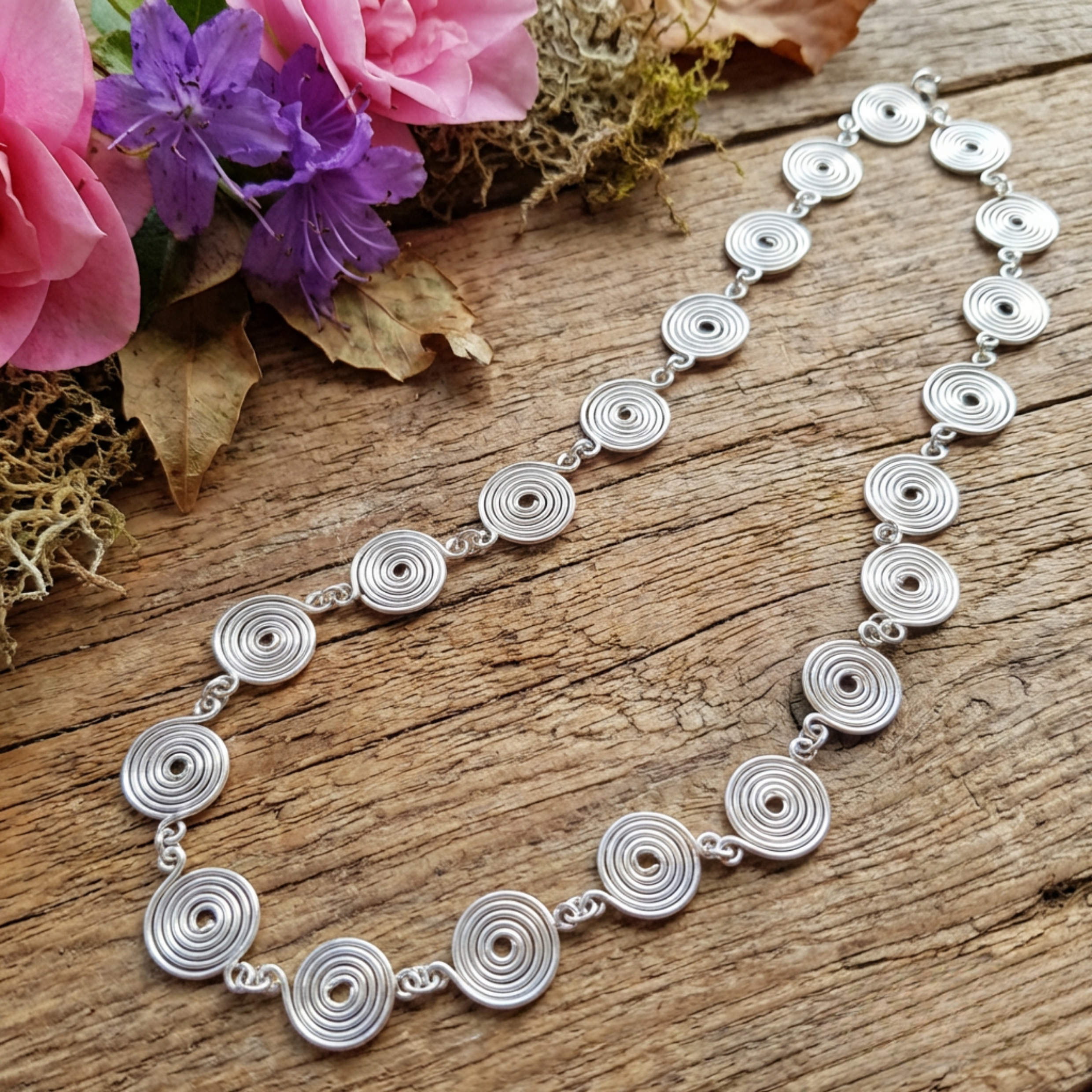 Silver spiral necklace with circular patterns on a wooden surface with flowers.