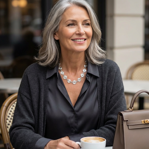 Woman sitting outdoors with a cup of coffee, wearing a grey cardigan and black shirt. And a closed silver spiral necklace.