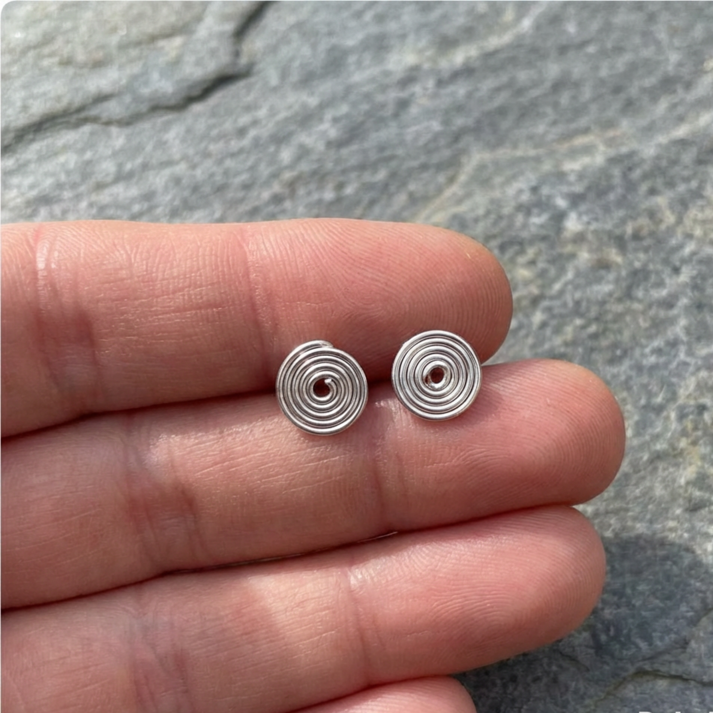 Silver spiral earrings held between fingers against a stone background
