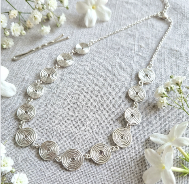 Silver necklace with circular design and half chain on a textured surface with white flowers
