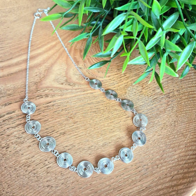 Overhead view of a silver spiral necklace with half chain on a wooden background with green leaves