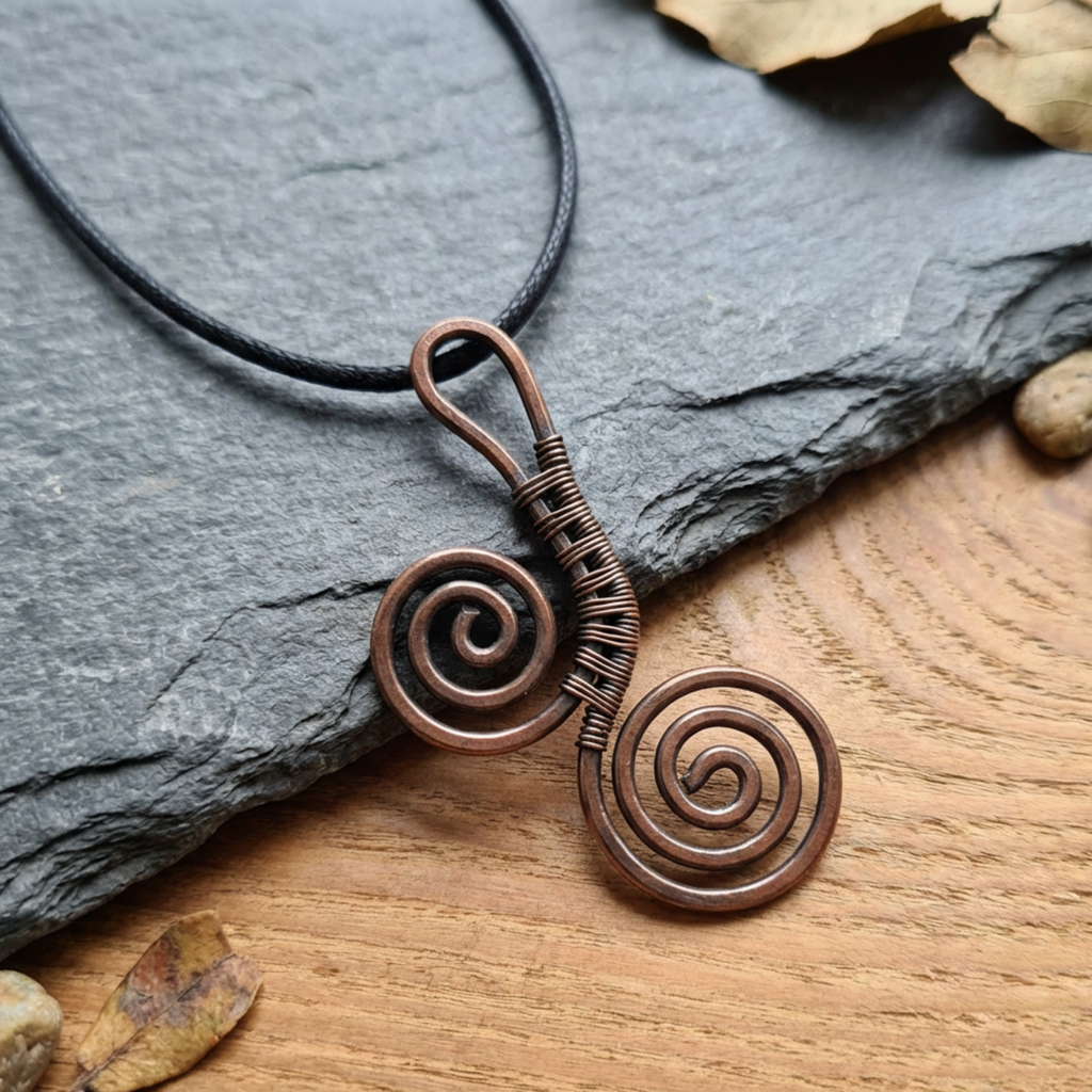 Copper spiral pendant necklace on a black cord, placed on a stone and wooden surface.