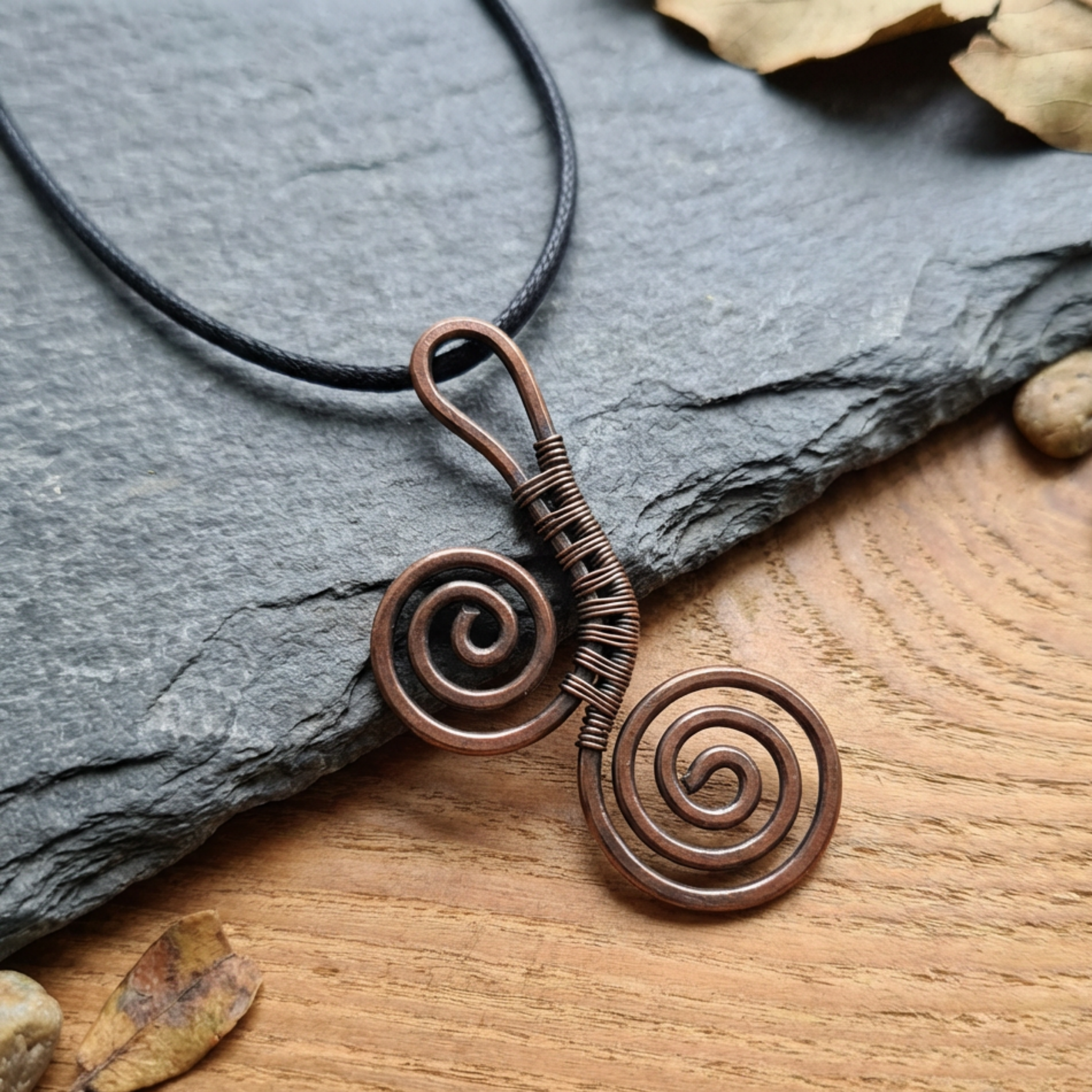 Copper spiral pendant necklace on a black cord, placed on a stone and wooden surface.