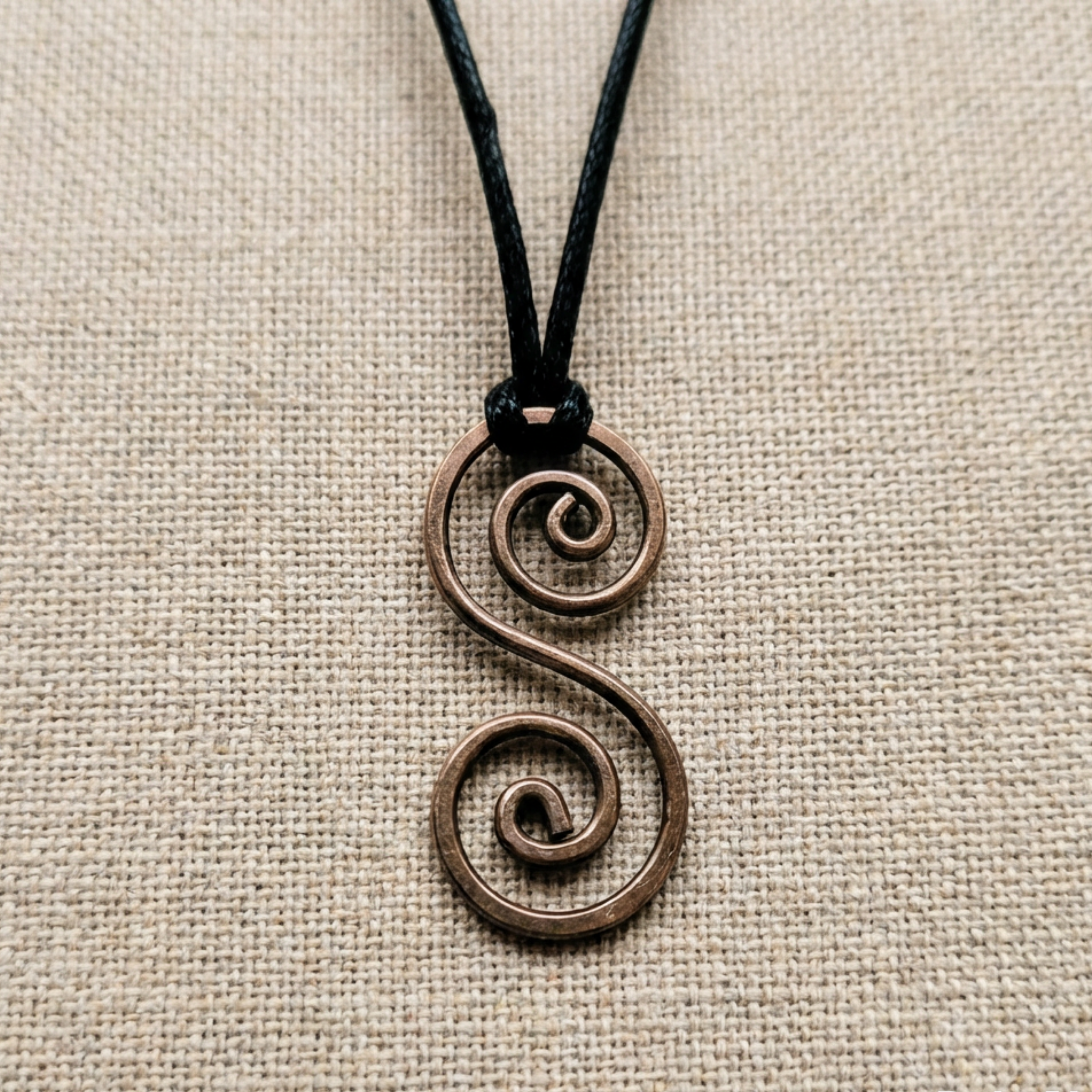 Bronze copper spiral pendant on a black cord against a beige fabric background