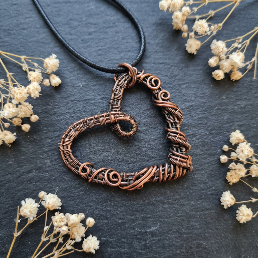Copper wire-wrapped heart-shaped pendant on a dark surface with small white flowers.