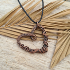 Copper heart-shaped pendant on a black cord against a wooden background with dried leaves.