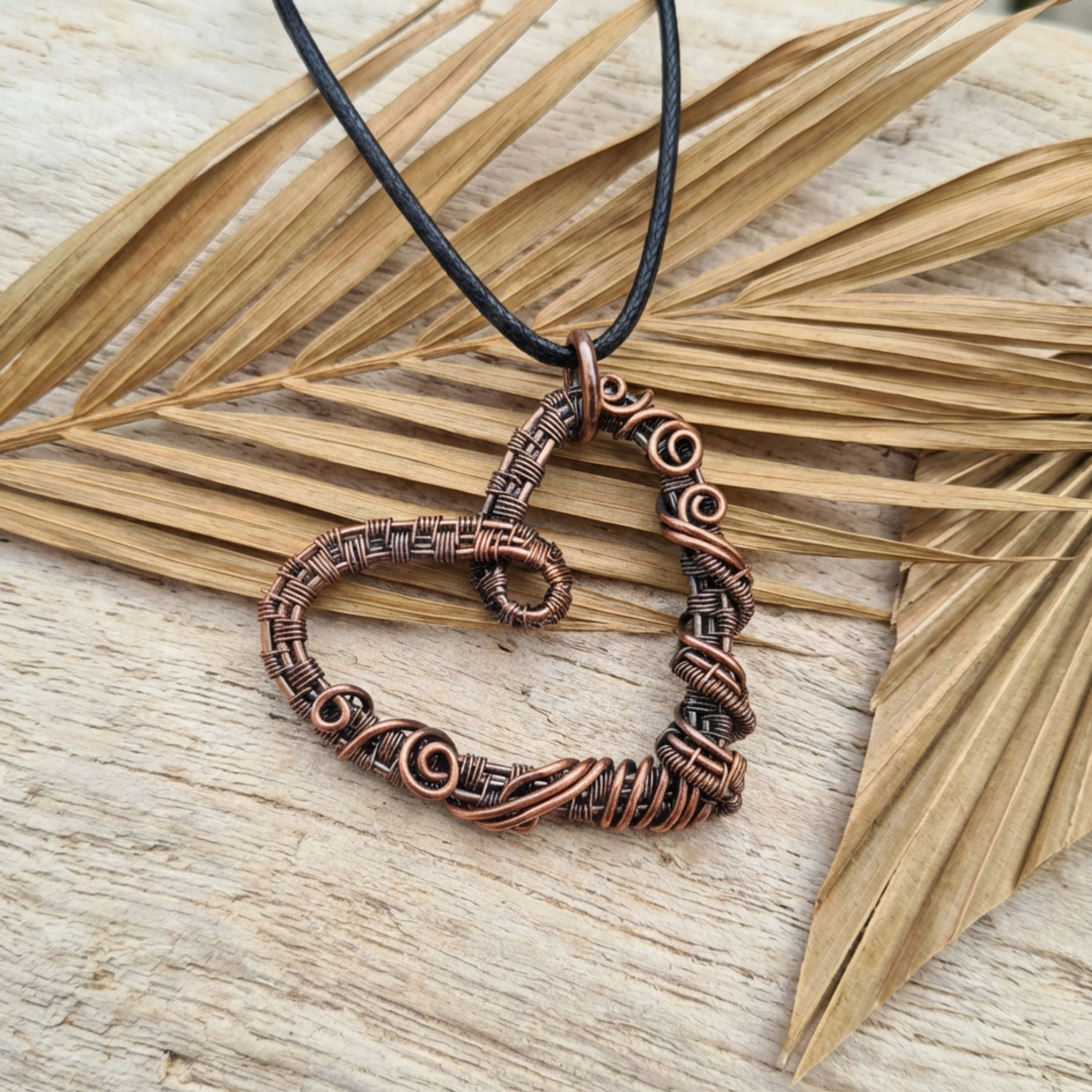 Copper heart-shaped pendant on a black cord against a wooden background with dried leaves.