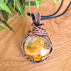 Copper-wrapped pendant with a natural stone on a wooden surface with green leaves in the background