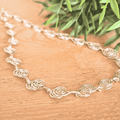Silver spiral necklace with floral patterns on a wooden surface with green leaves in the background