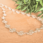 Silver spiral necklace with floral patterns on a wooden surface with green leaves in the background