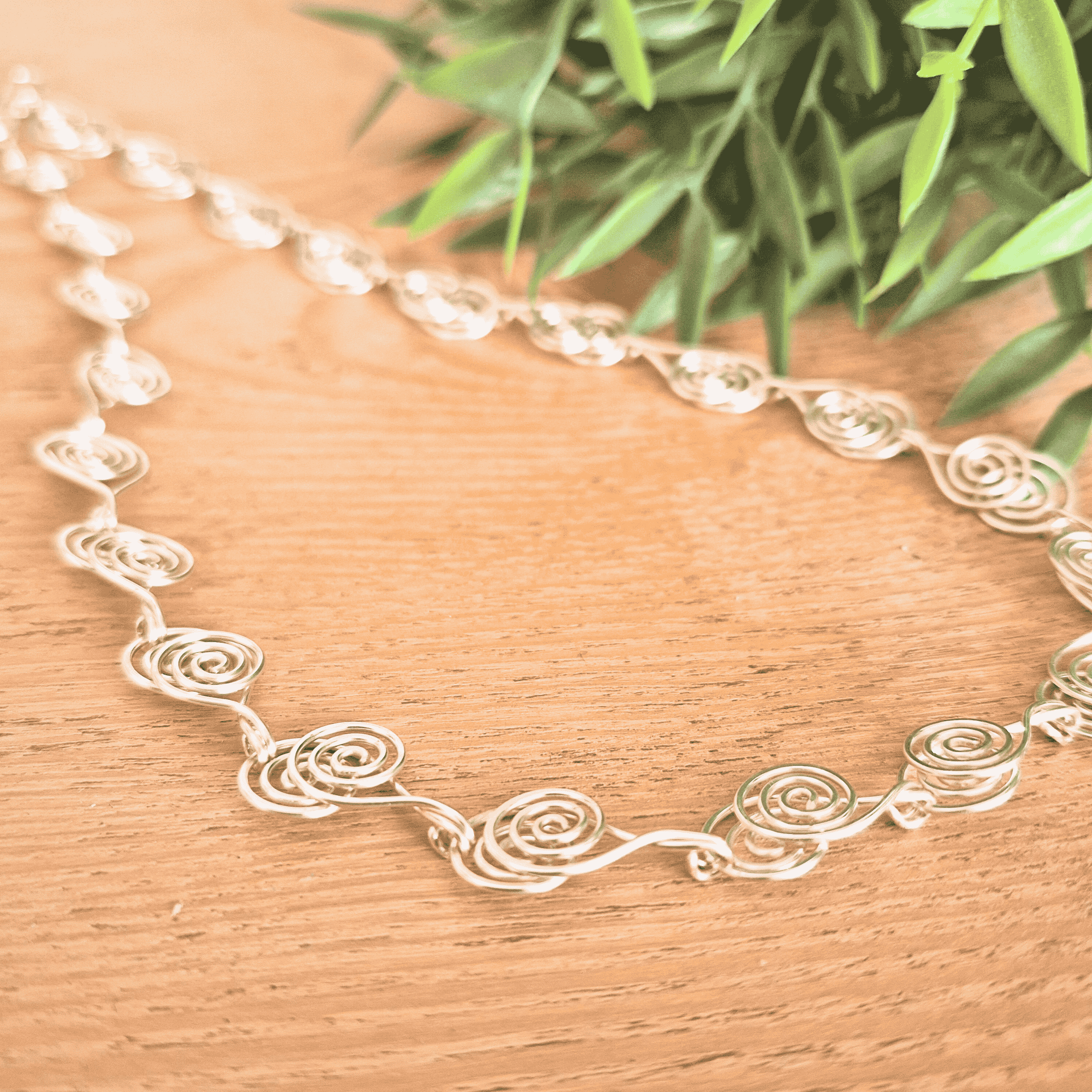 Silver spiral necklace with floral patterns on a wooden surface with green leaves in the background