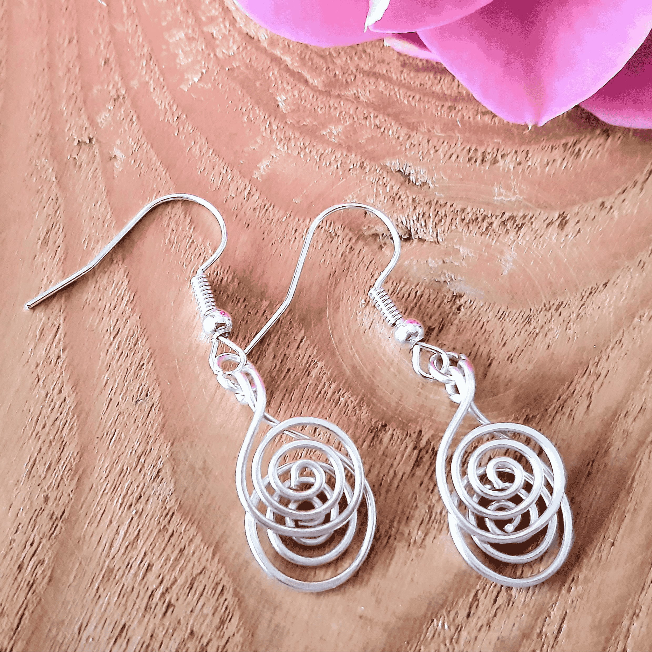 Silver spiral earrings on a wooden surface with pink flowers in the background