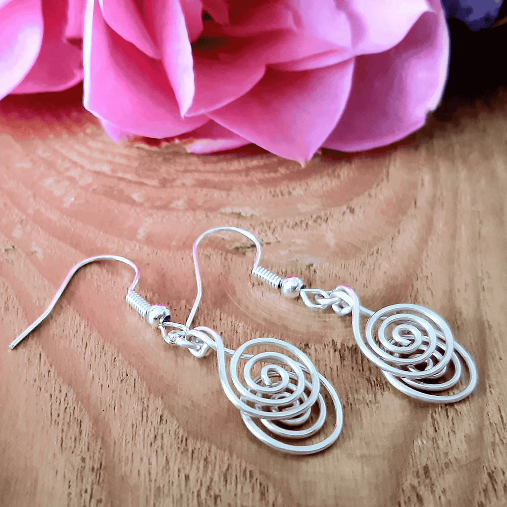 Silver spiral earrings on a wooden surface with pink flowers in the background