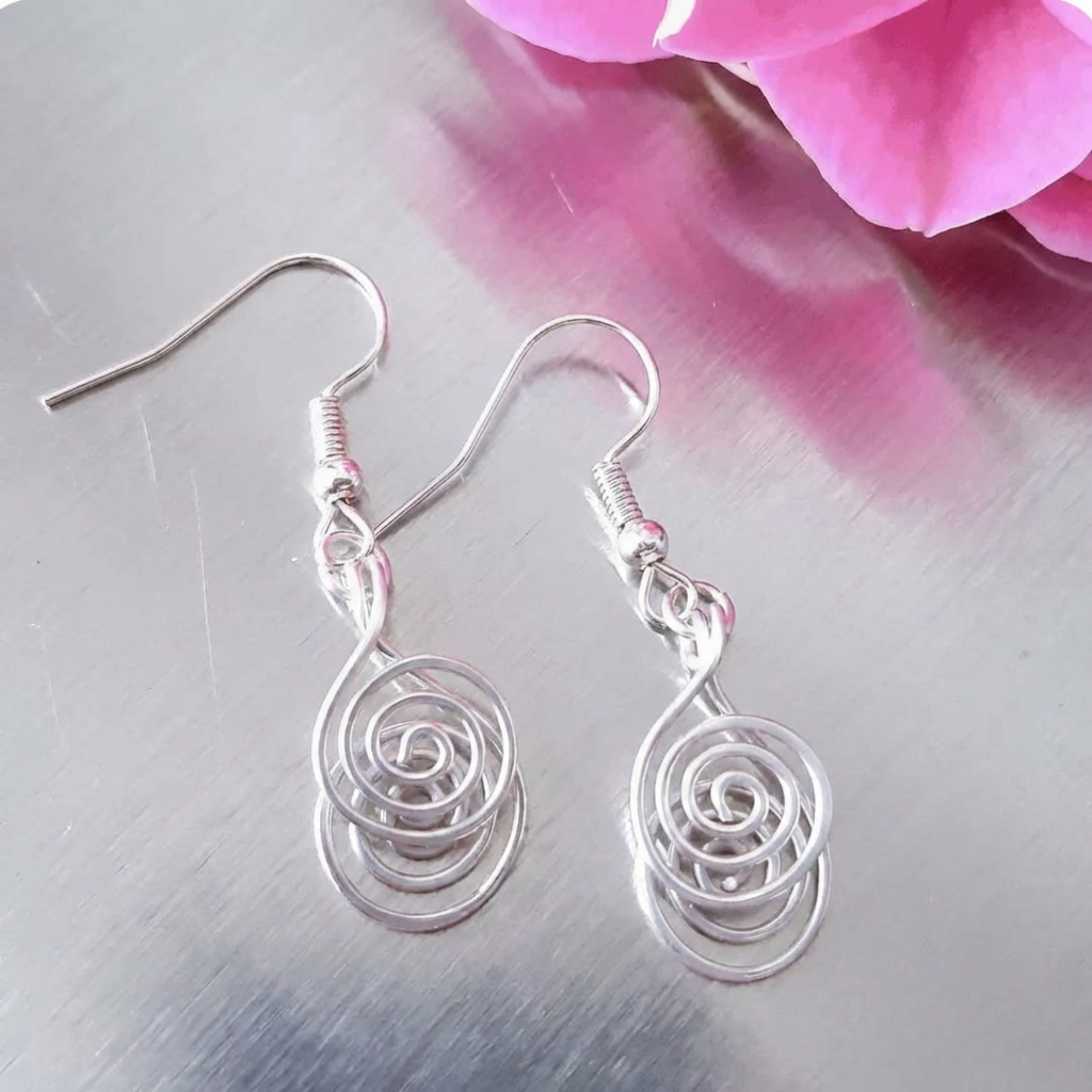 Silver spiral earrings on a metallic surface with pink flowers in the background