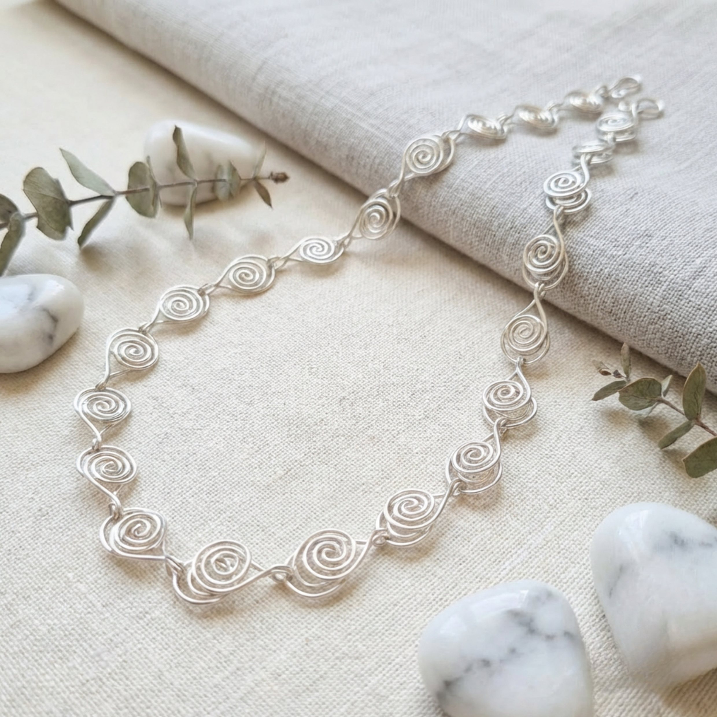 Silver spiral necklace on a textured surface with marble stones and greenery