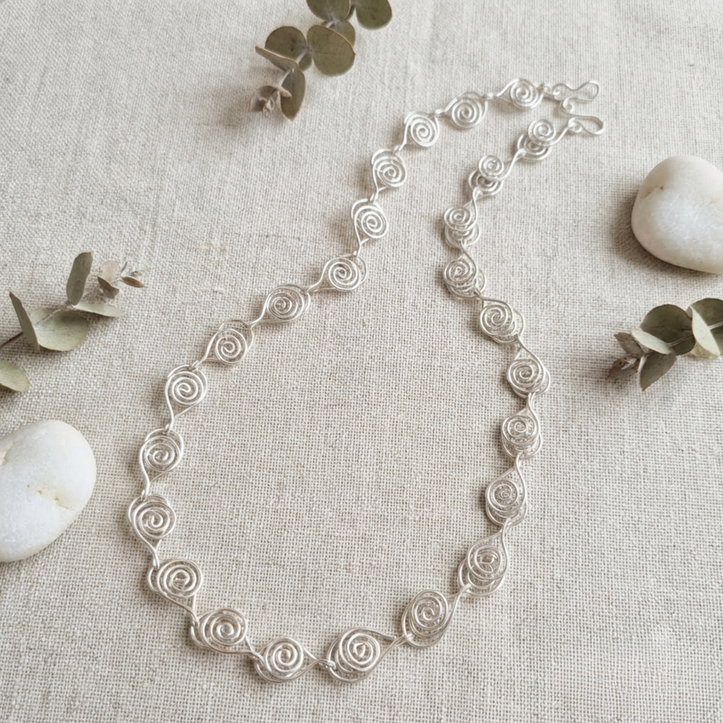 Silver necklace on a textured beige surface with eucalyptus leaves and stones.