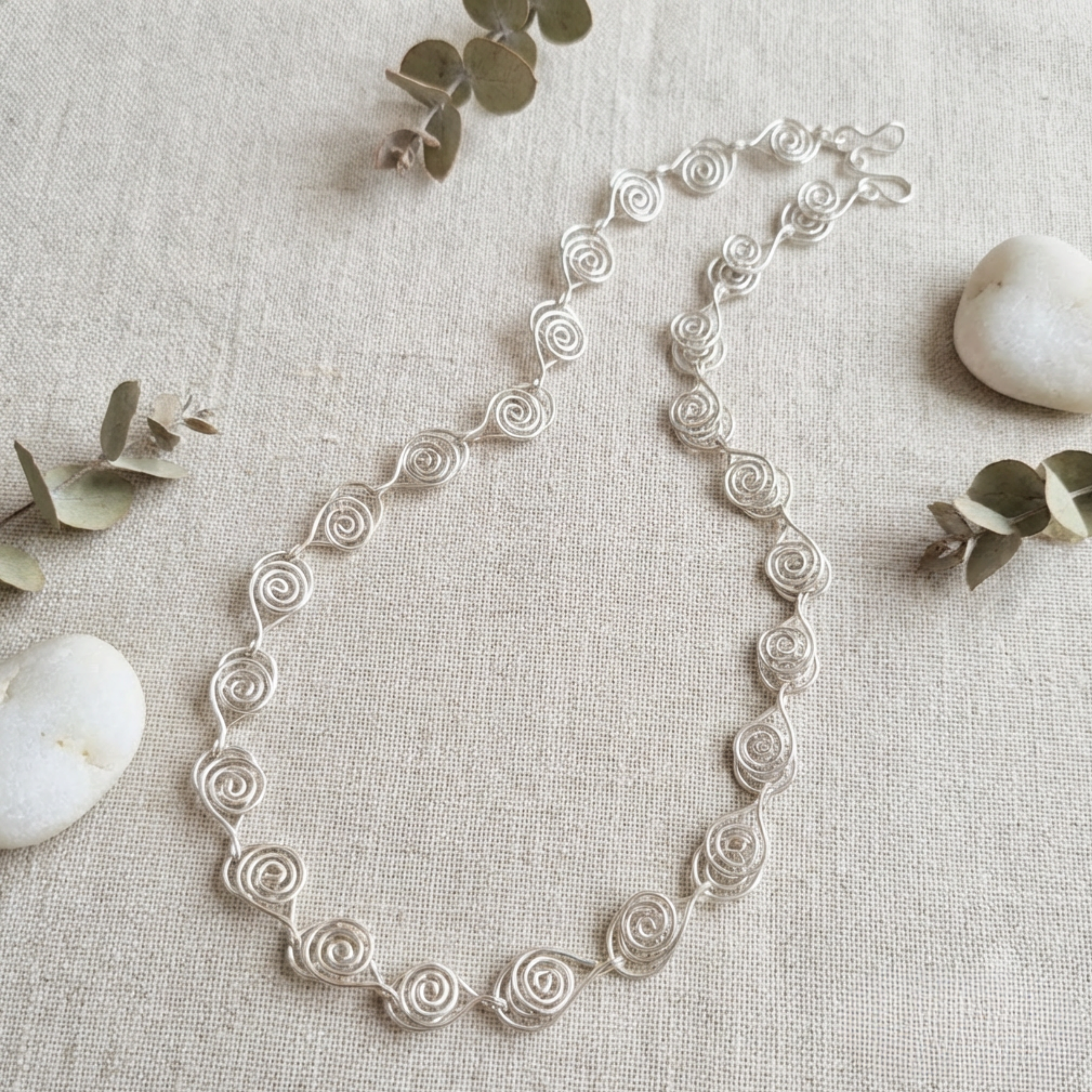 Silver necklace on a textured beige surface with eucalyptus leaves and stones.